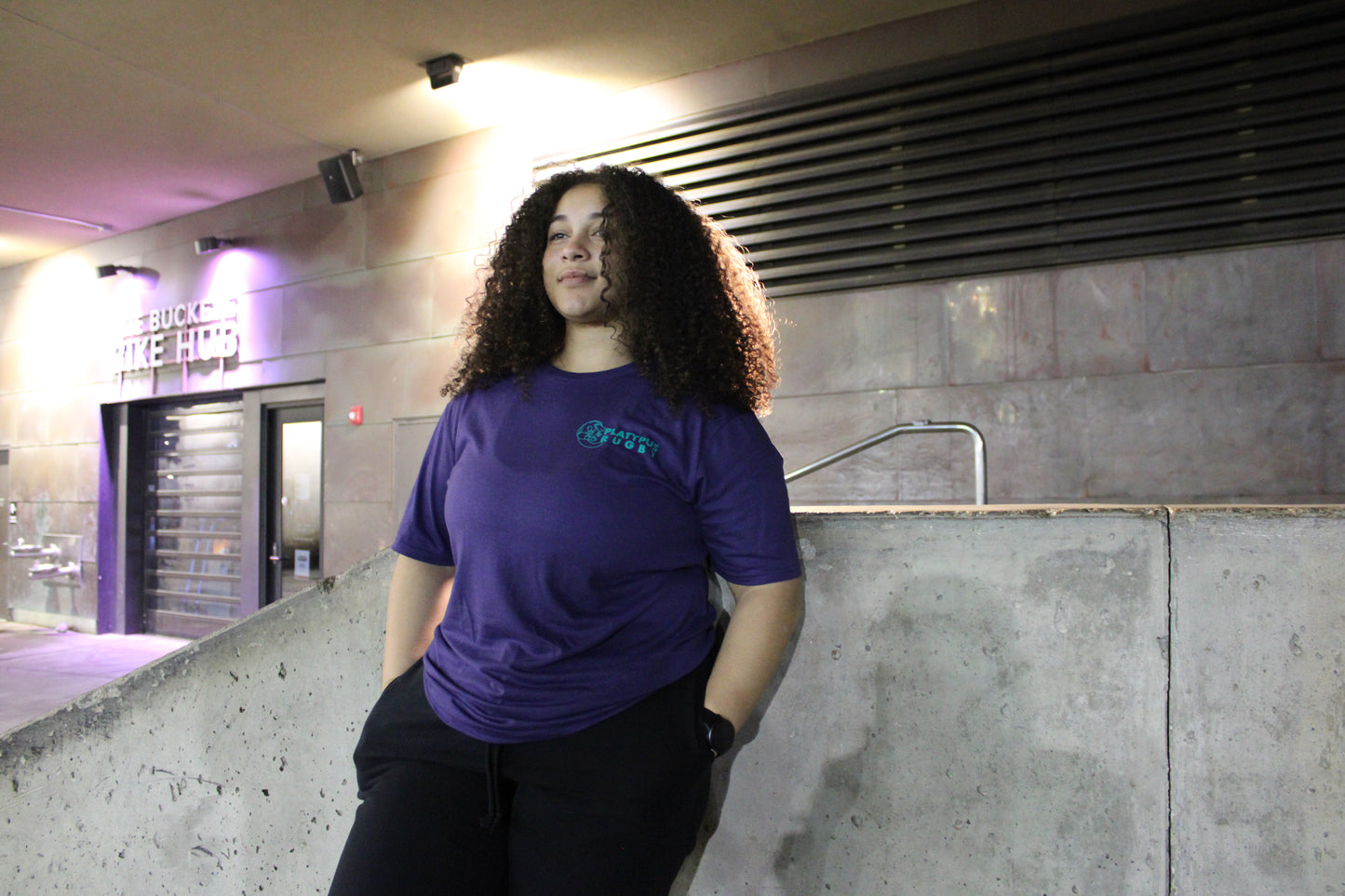 Person wearing a dark blue shirt standing in an indoor setting with concrete walls and a staircase.