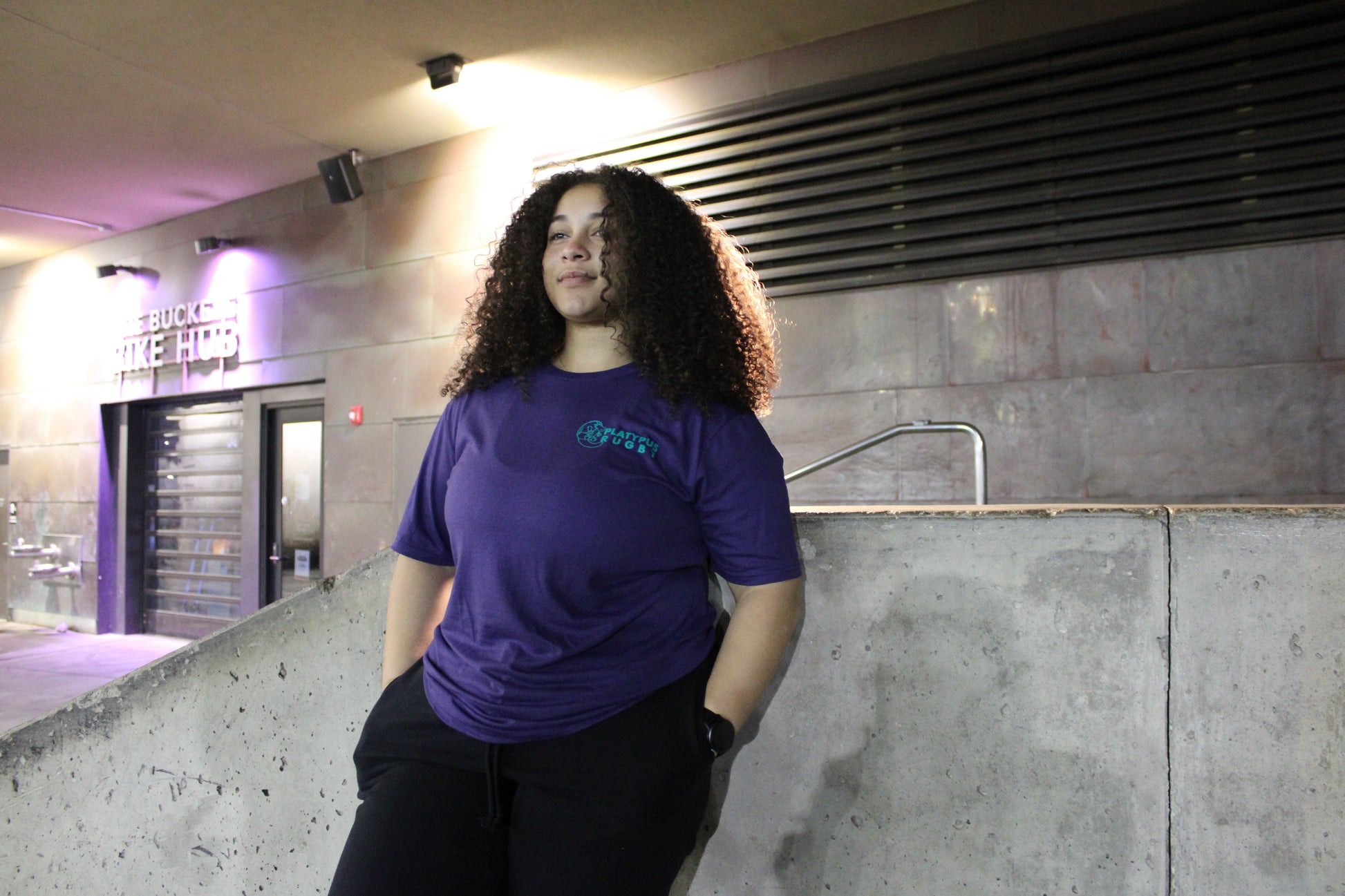 Person wearing a dark blue shirt standing in an indoor setting with concrete walls and a staircase.